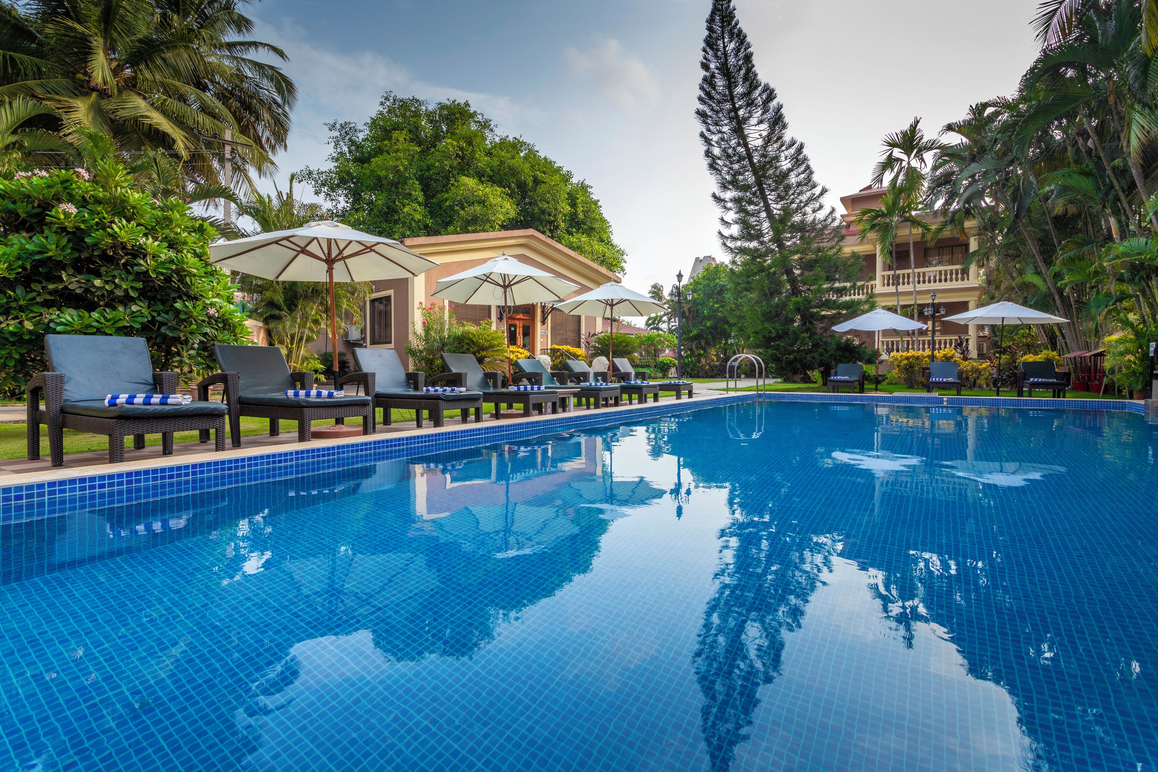 A swimming pool with lounge chairs and umbrellas, surrounded by lush greenery and a building in the background.