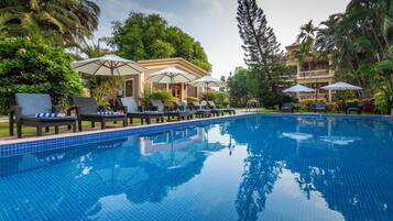 A swimming pool with lounge chairs and umbrellas, surrounded by lush greenery and a building in the background.