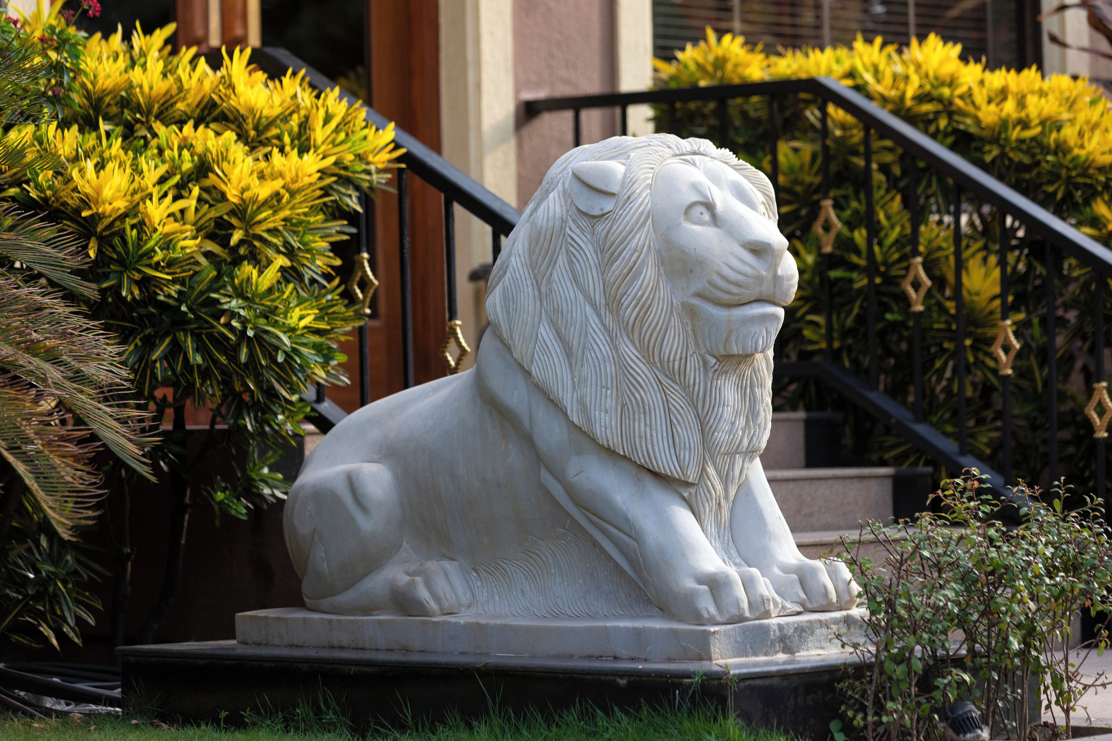 Une statue de lion en pierre, située dans un jardin aménagé, avec un escalier et de la verdure en arrière-plan.