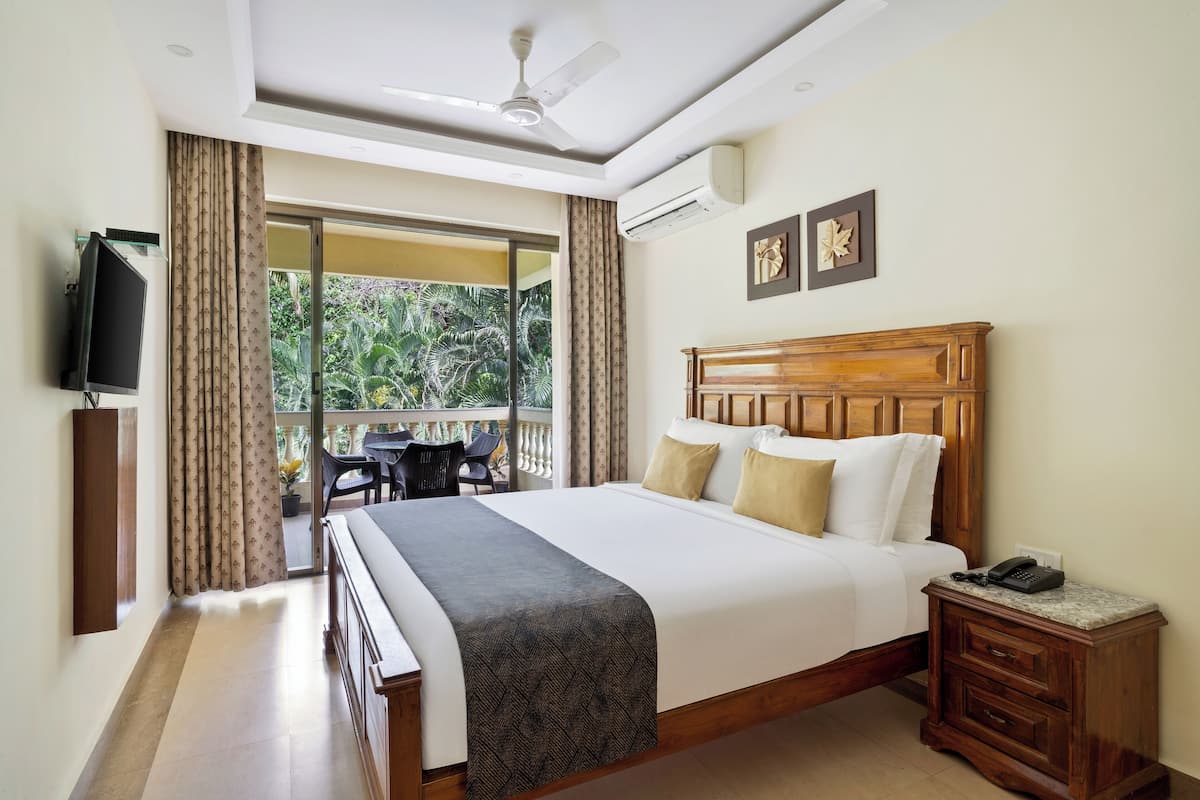 A hotel room with a large bed, a wooden headboard, a bedside table with a phone, and a view of a balcony with outdoor furniture.