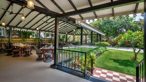 Un espace de restauration intérieur avec des tables et des chaises, une balustrade en verre donnant sur un jardin et un motif de carreaux sur le sol.