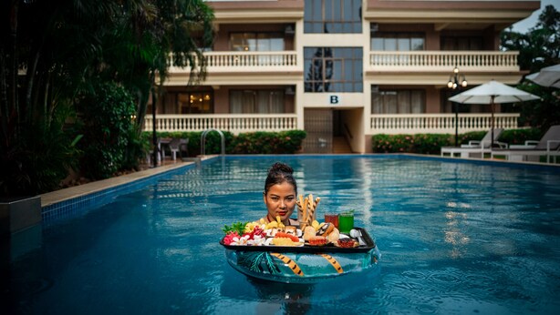 Une vue sur la piscine, avec un espace repas aménagé sur un bateau, un bâtiment doté de balcons et une végétation luxuriante.