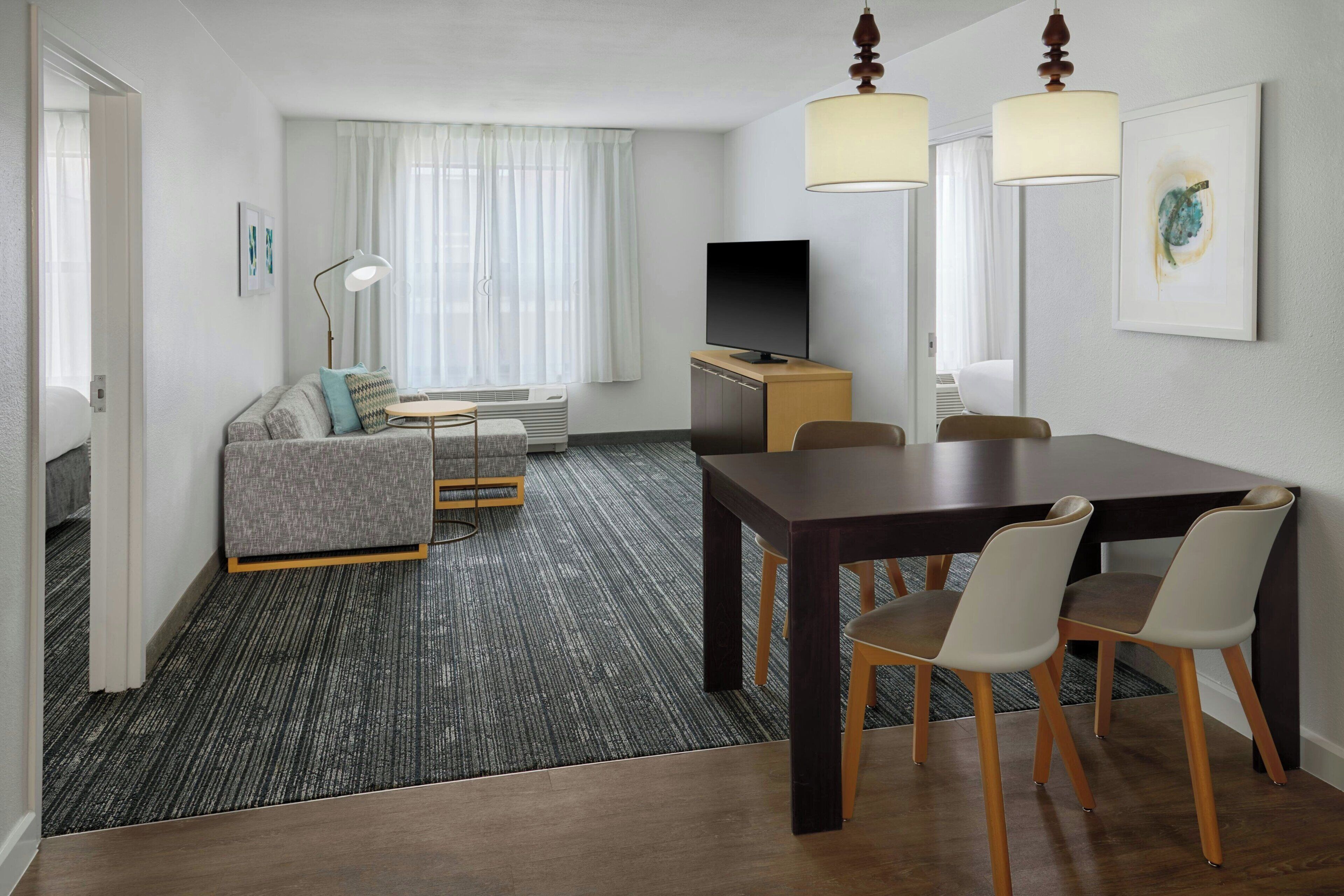 Quarto de hotel com sofá, televisão, mesa de jantar com cadeiras e uma janela grande com cortinas.