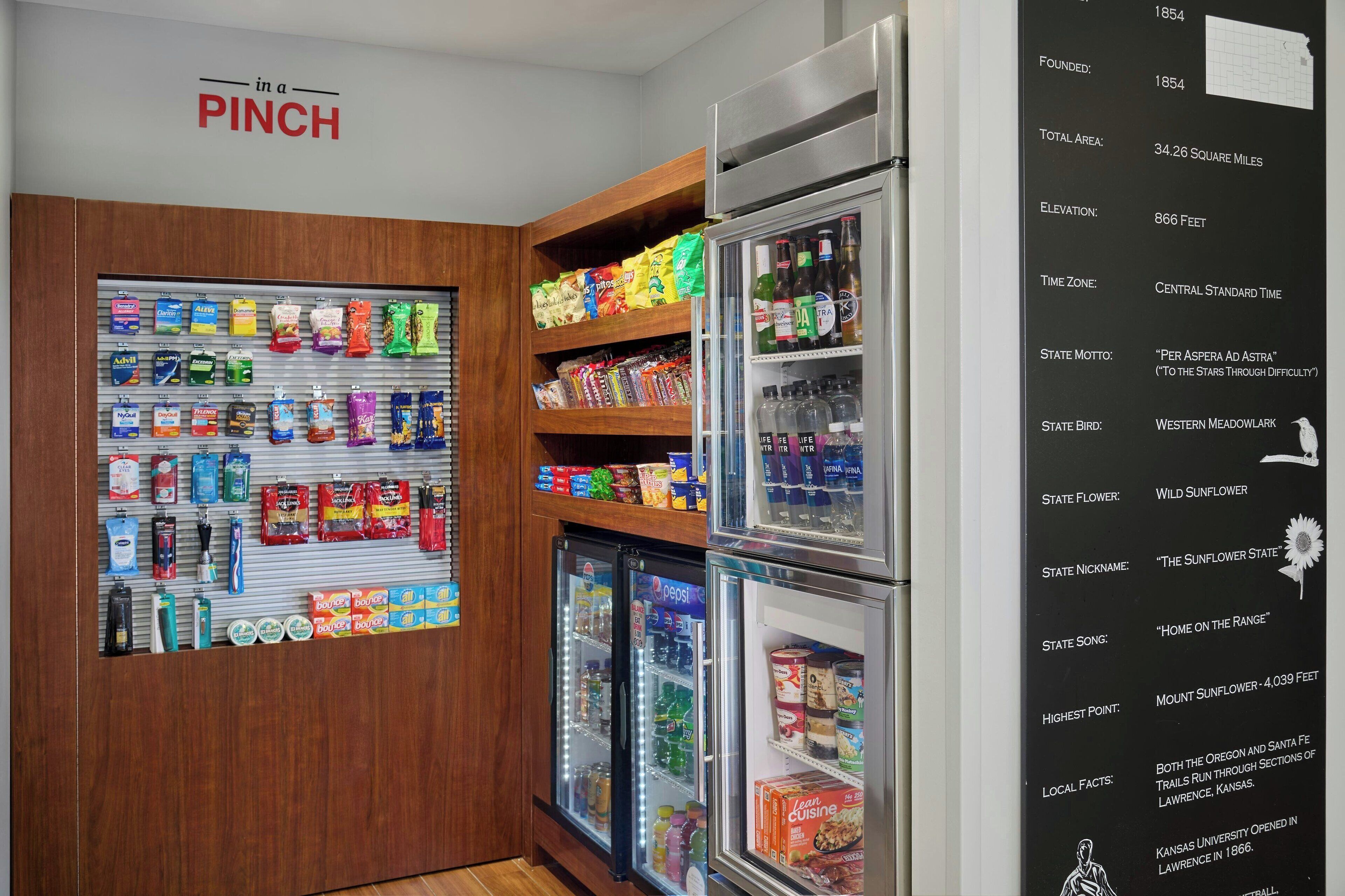 A snack display with various packaged foods and beverages, a refrigerator with drinks, and a wall with a list of facts about a location.