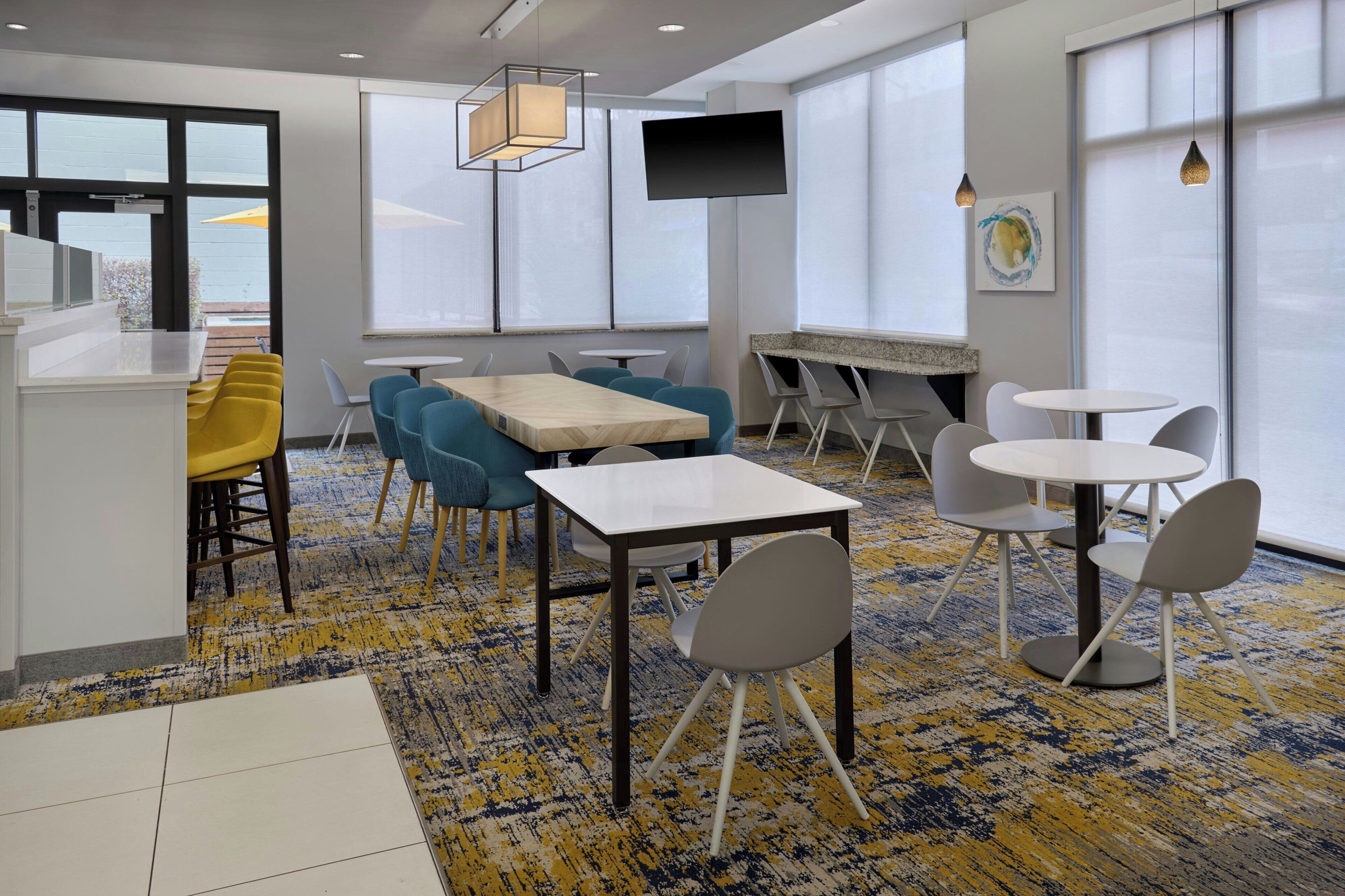 Una habitación de hotel moderna con un ventanal grande, un televisor de pantalla plana, un comedor con mesa y sillas, y suelo alfombrado.