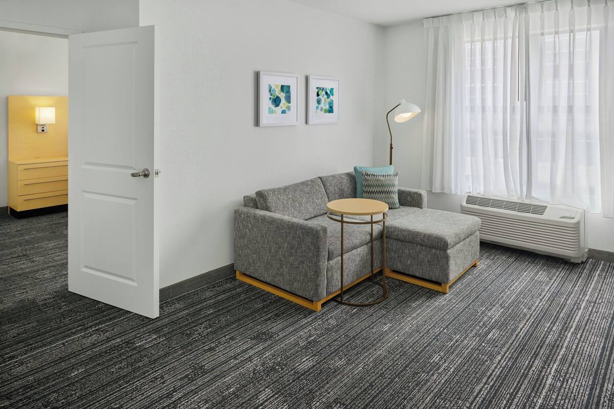 A modern hotel room with a grey sofa, a small round table, and a large window with sheer curtains.