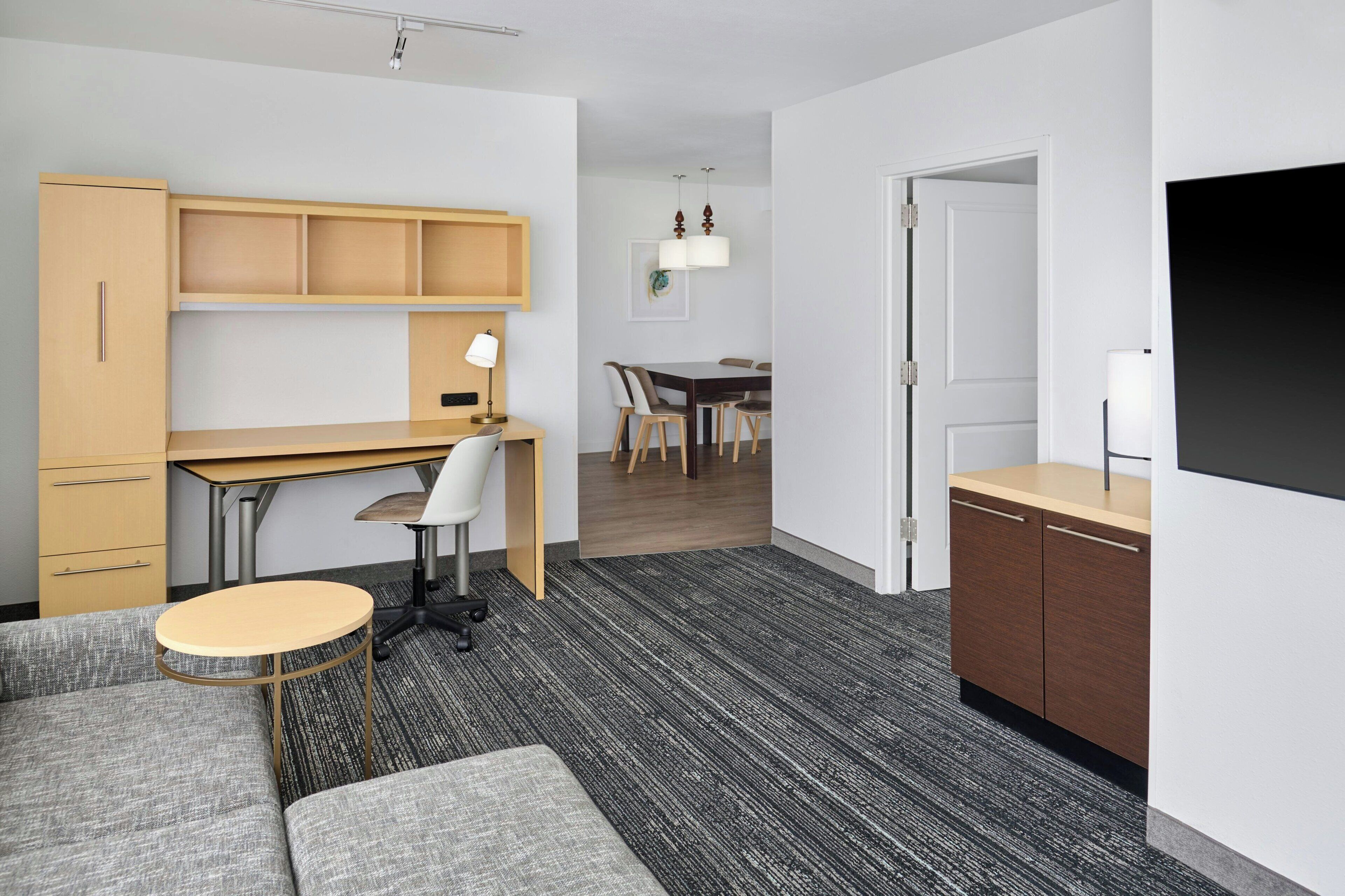 Et moderne hotelværelse med sofa, et lille rundt bord, et skrivebord med stol, et spiseområde med bord og stole, samt et fjernsyn monteret på væggen.