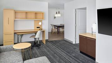 Et moderne hotelværelse med sofa, et lille rundt bord, et skrivebord med stol, et spiseområde med bord og stole, samt et fjernsyn monteret på væggen.