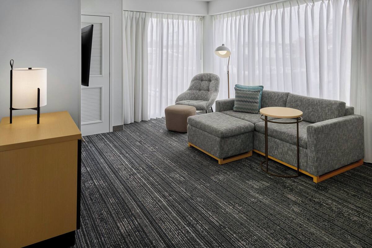 A modern hotel room with a grey sofa, a small round table, a lamp, and a TV mounted on the wall.