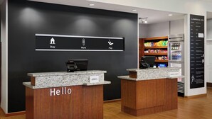 Uma área de recepção de hotel com dois balcões identificados com “Hello” e “Marriott Bonvoy”, uma parede preta com ícones para “Stay”, “Work” e “Play”, e uma prateleira com petiscos e bebidas.