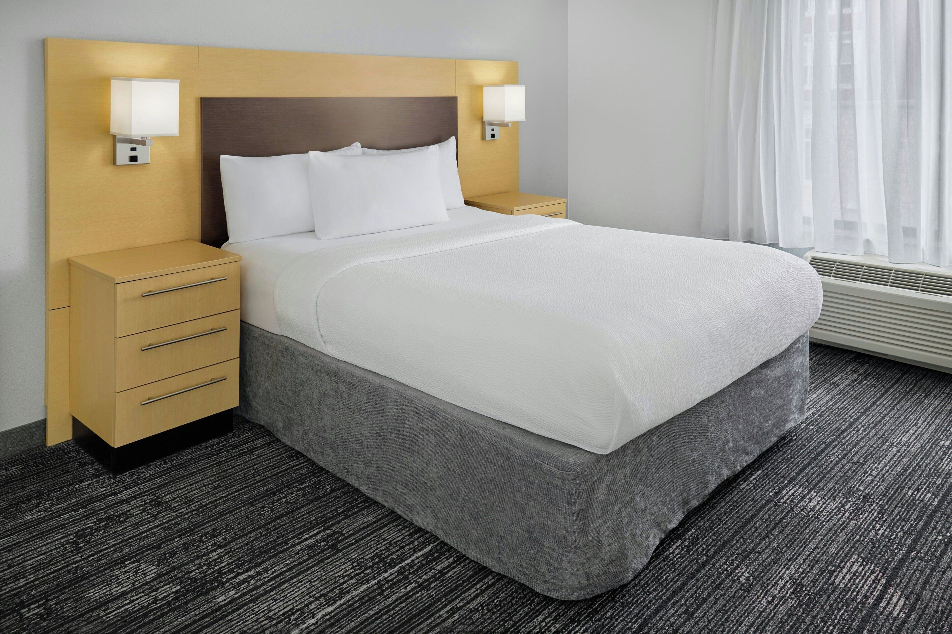 A hotel room with a large bed, two bedside tables, a nightstand, a wall-mounted lamp, and a window with sheer curtains.
