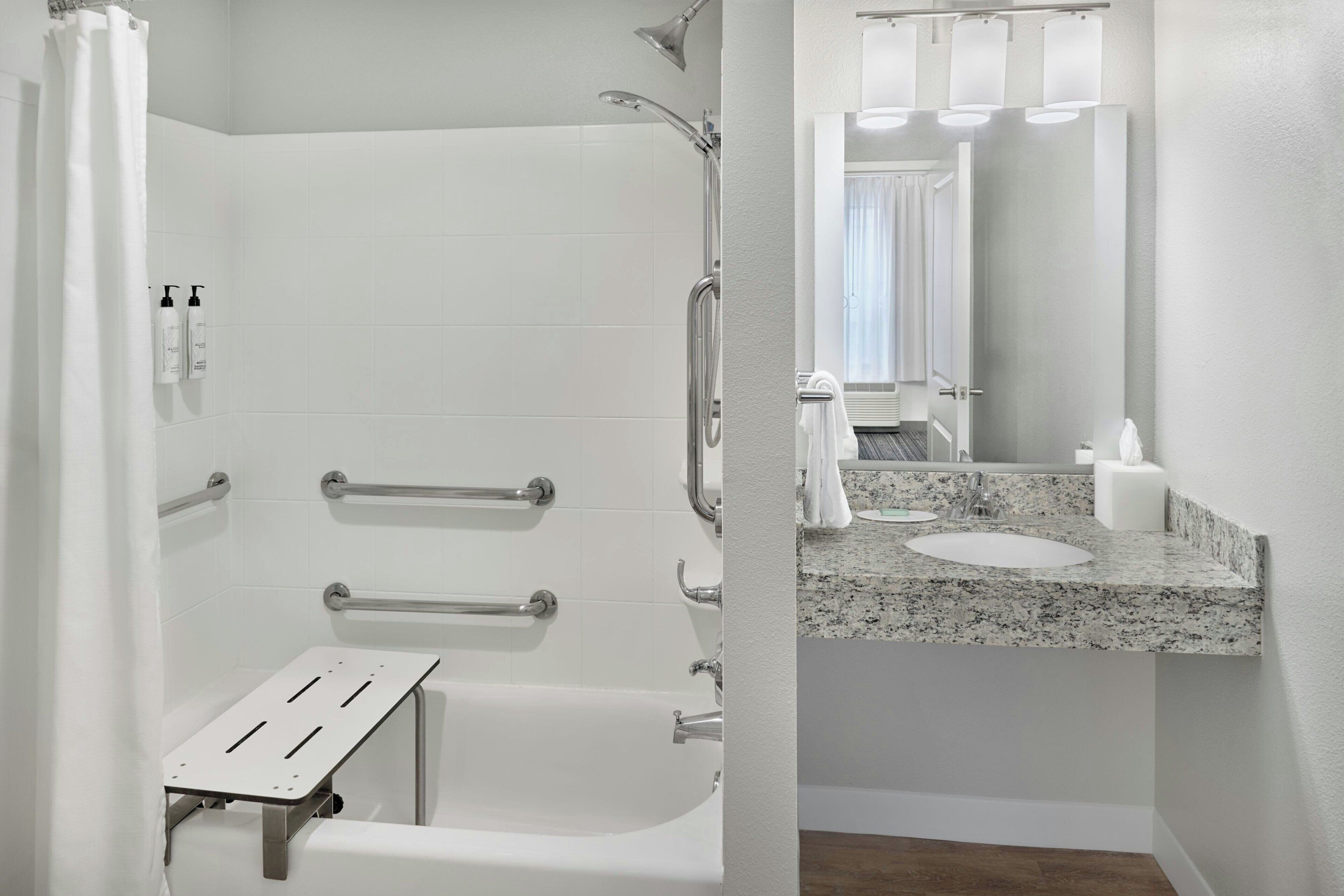 A bathroom with a shower, grab bars, a white bathtub, a granite countertop sink, and a mirror.