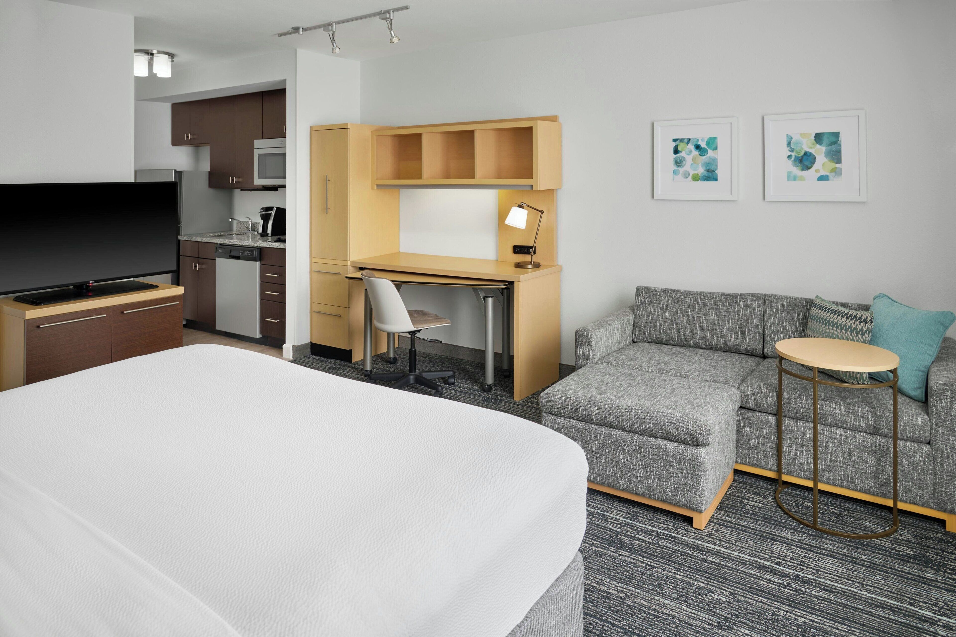 Een moderne hotelkamer met een groot bed, een grijze bank, een klein bureau en een kitchenette.