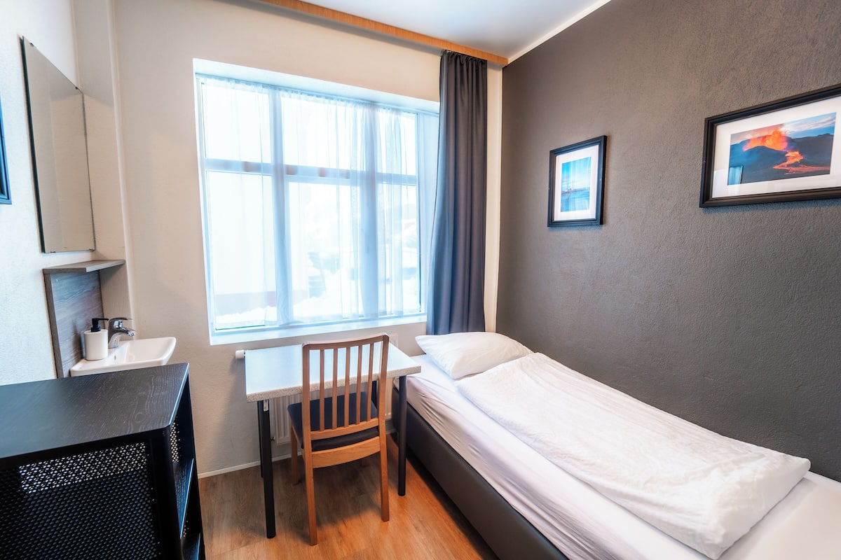 A room with a bed, a desk, a chair, a window with curtains, and two framed pictures on the wall.