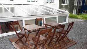 A wooden table and chairs set on a gravel surface, with a glass-enclosed patio area in the background.