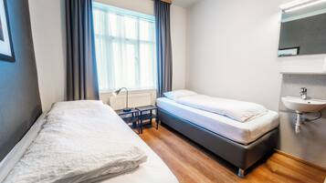 A twin room with a bed, a bedside table, a window with curtains, a radiator, a wall-mounted sink, and a mirror.