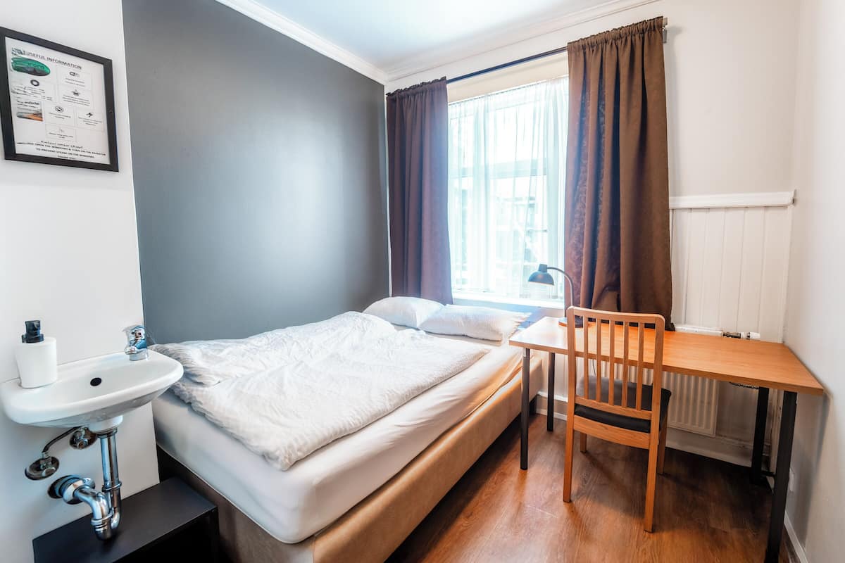 A bedroom with a bed, a wooden table, a chair, a white sink, and a window with curtains.