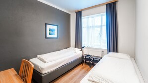 A hotel room with two single beds, a wooden table, a chair, a desk with a lamp, a framed picture on the wall, and a window with curtains.