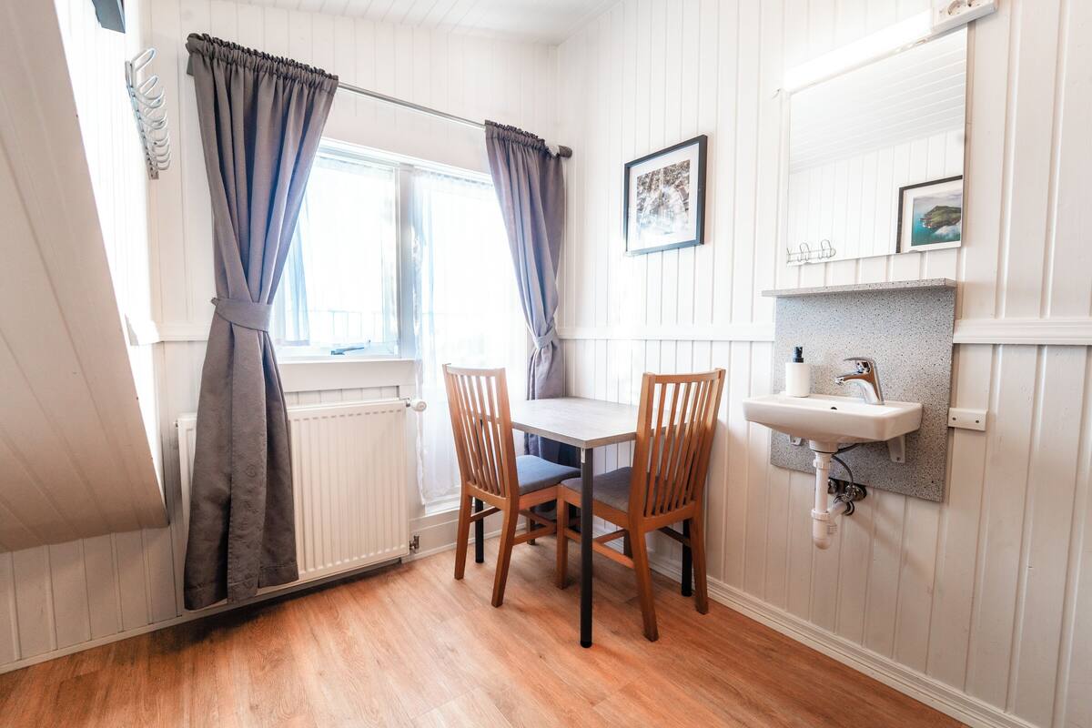 A room with a wooden table, two chairs, a window with curtains, a wall-mounted sink, and a framed picture.
