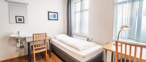 Une petite pièce propre, avec un lit, un bureau, une chaise, un lavabo et une fenêtre avec des rideaux.