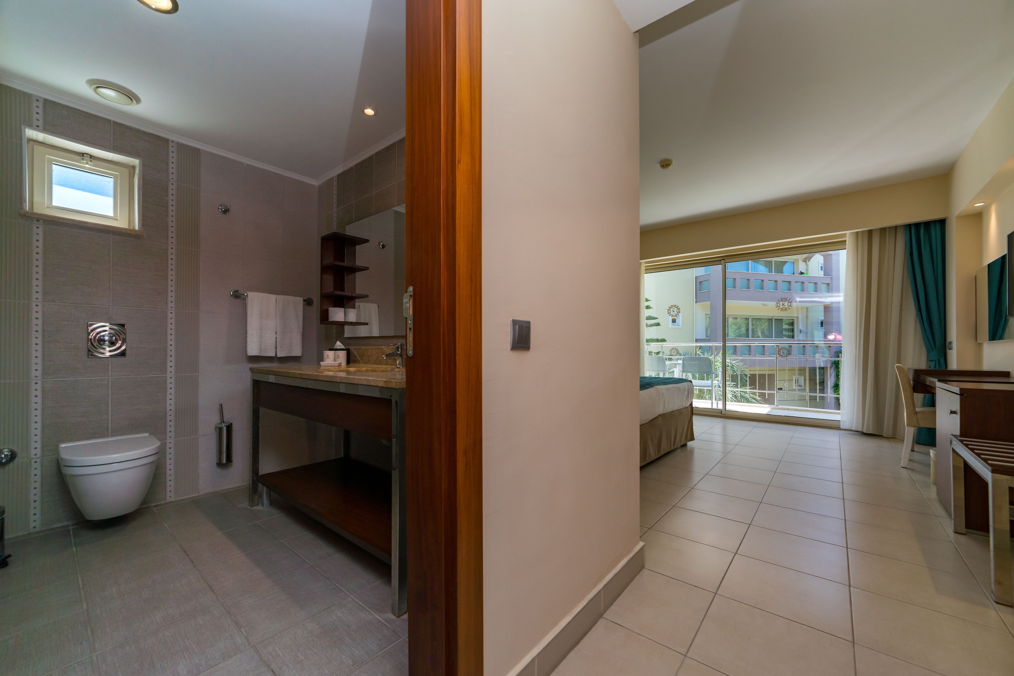 A modern bathroom with a toilet, sink, and vanity, a wooden door leading to a balcony with a view of buildings.