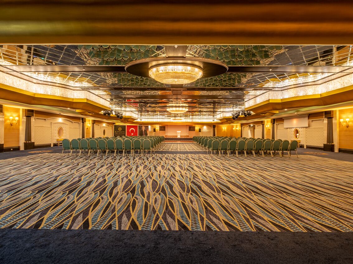 Una amplia sala de conferencias con filas de sillas mirando hacia un escenario, una alfombra estampada y un techo decorativo.