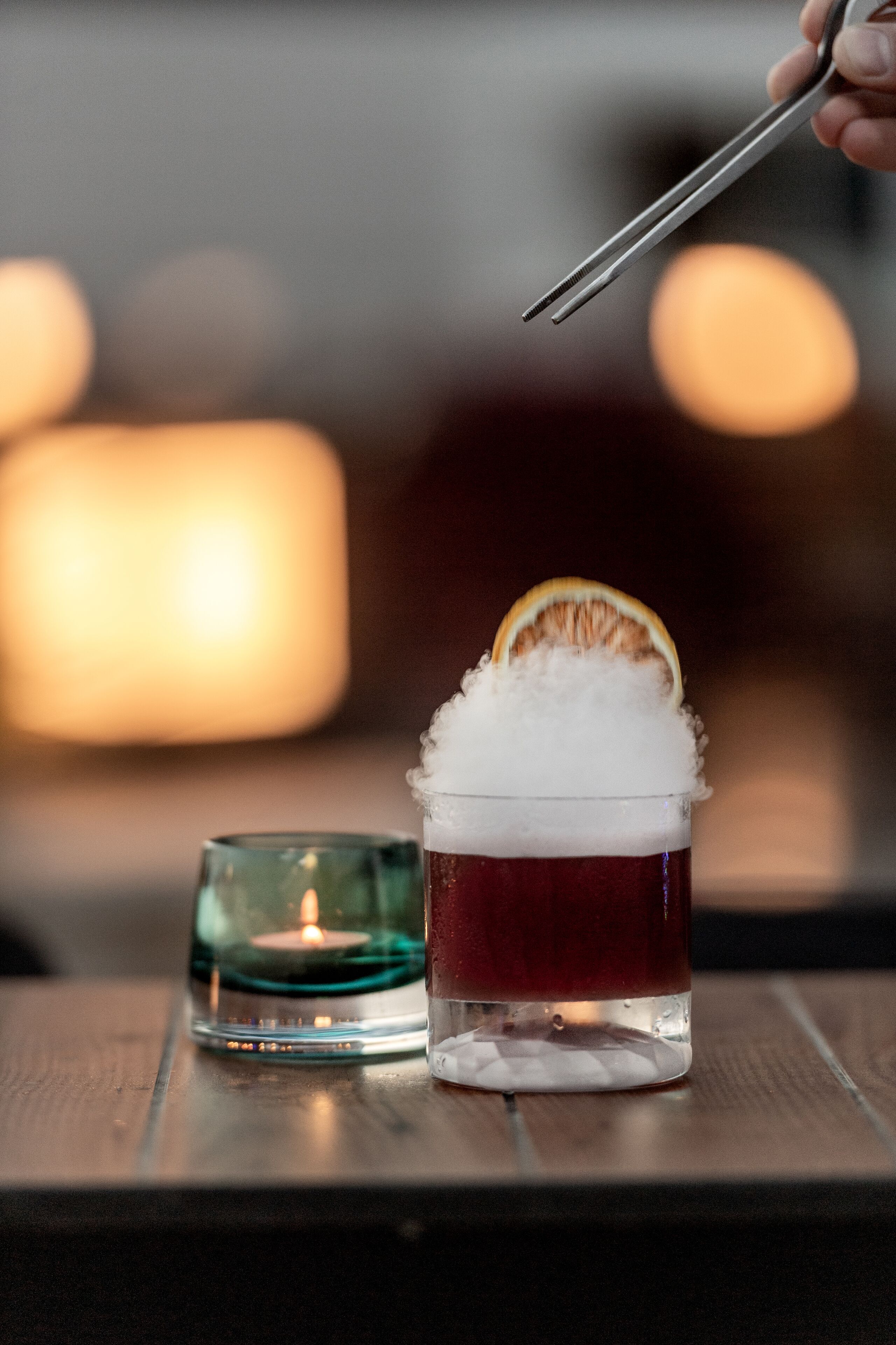 A glass with a red beverage and a slice of orange on top, placed on a wooden surface with a lit candle in the background.