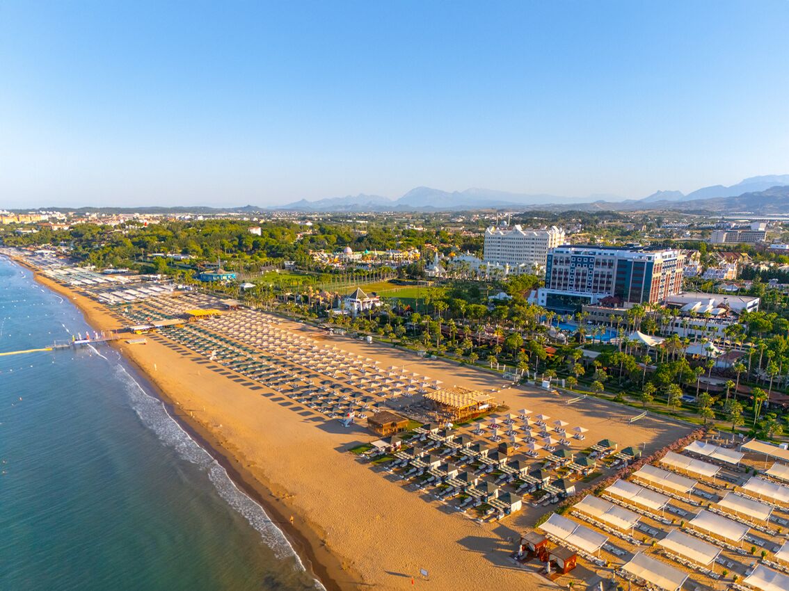 An aerial view of a beach resort with rows of sunbeds and umbrellas, a large building, and a clear view of the ocean and mountains in the distance.