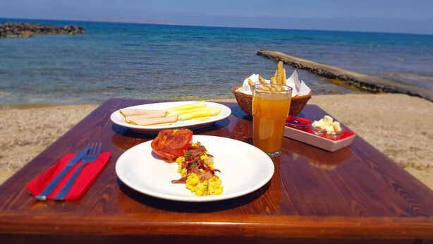 A wooden table with a meal consisting of scrambled eggs, a slice of meat, and bread, accompanied by a glass of iced tea, set on a beach with the sea in the background. - Haris Apartments (Hersonissos)