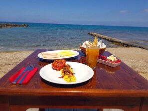 A wooden table with a meal consisting of scrambled eggs, a slice of meat, and bread, accompanied by a glass of iced tea, set on a beach with the sea in the background. - Haris Apartments (Hersonissos)