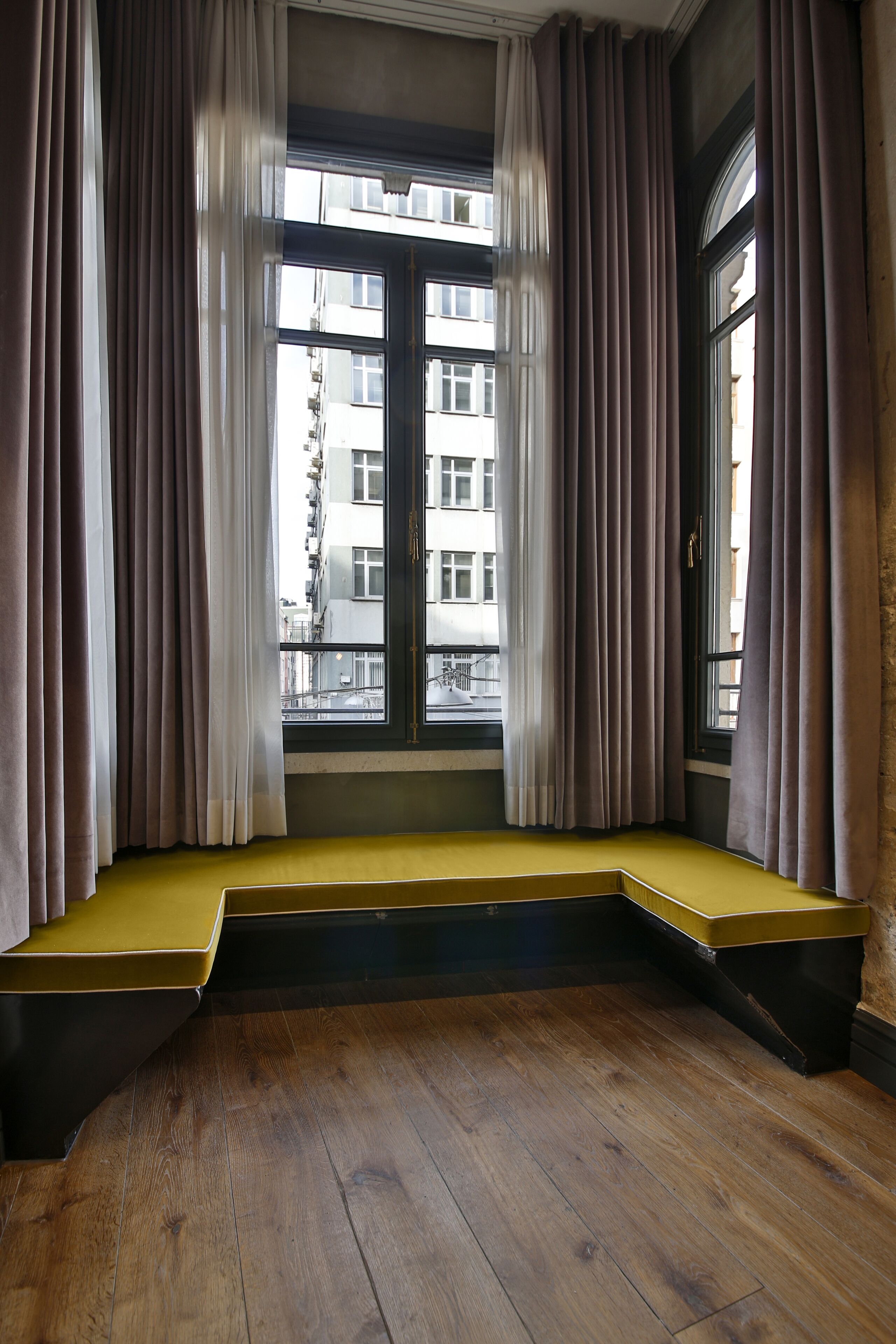 A room with a built-in bench seat, a window with curtains, and a view of buildings outside.