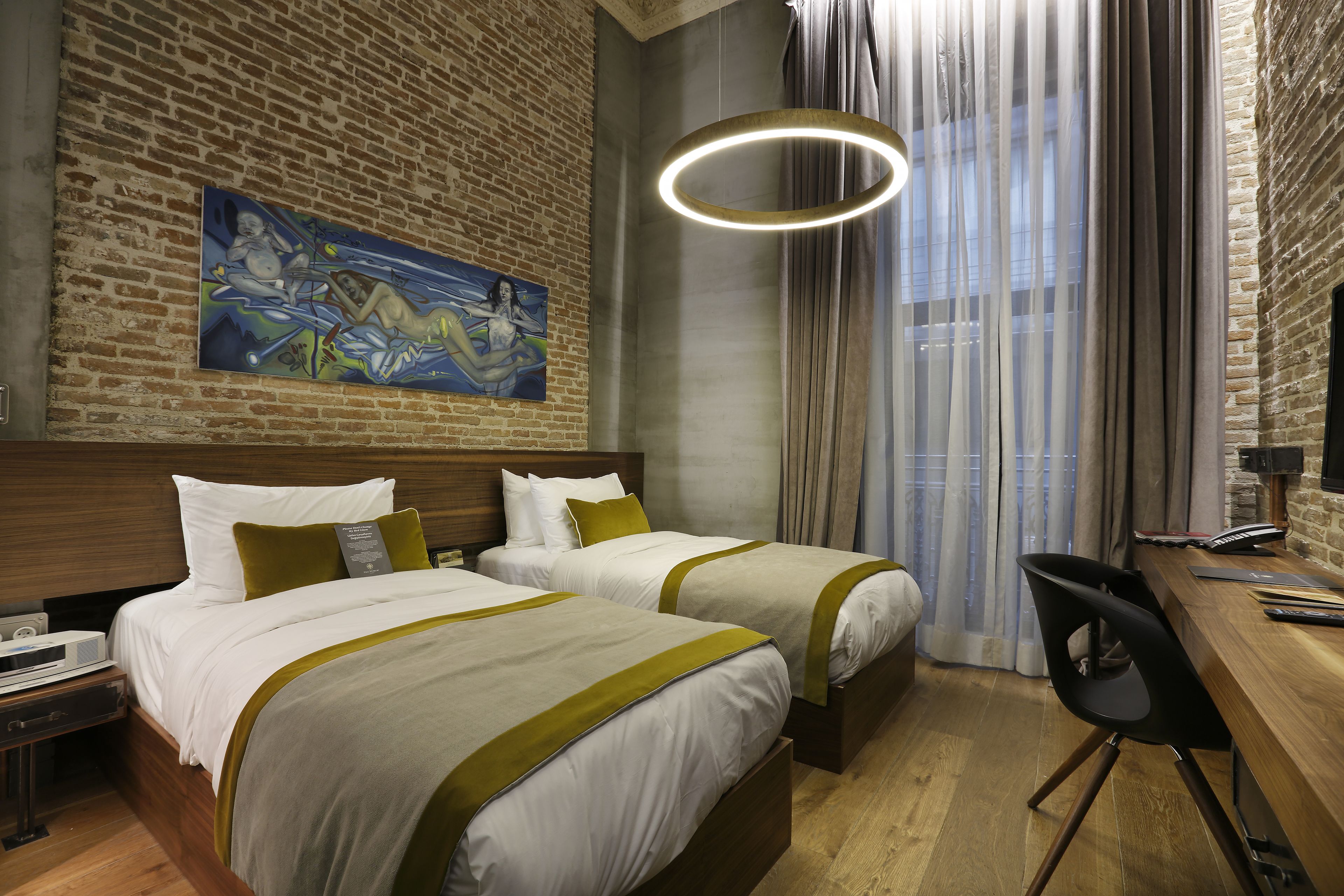 A hotel room with two beds, a desk, a chair, a painting on the wall, and a large window with curtains.
