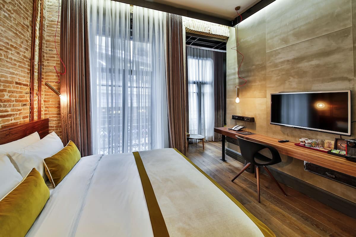Habitación de hotel con una cama grande, un escritorio con televisor y vistas a la ciudad a través de amplias ventanas.