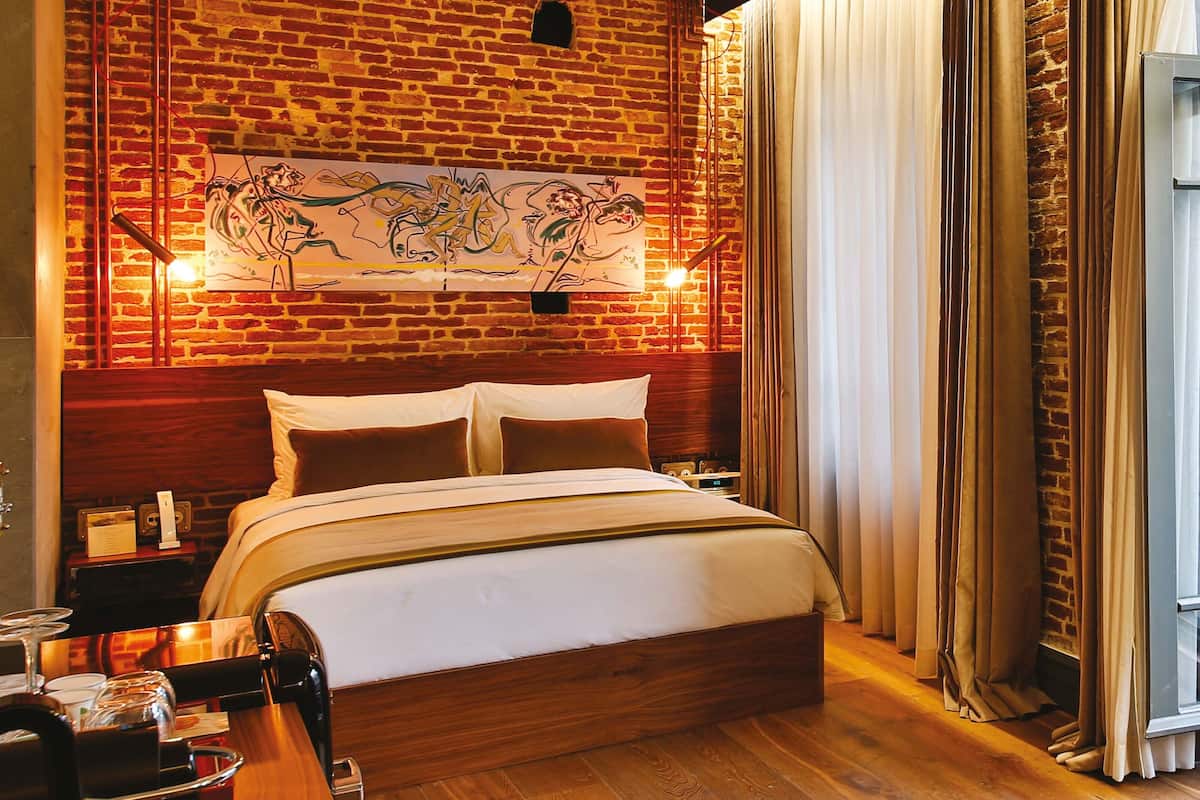 Una habitación de hotel con pared de ladrillo, una cama grande, un escritorio con cafetera y una ventana con cortinas.