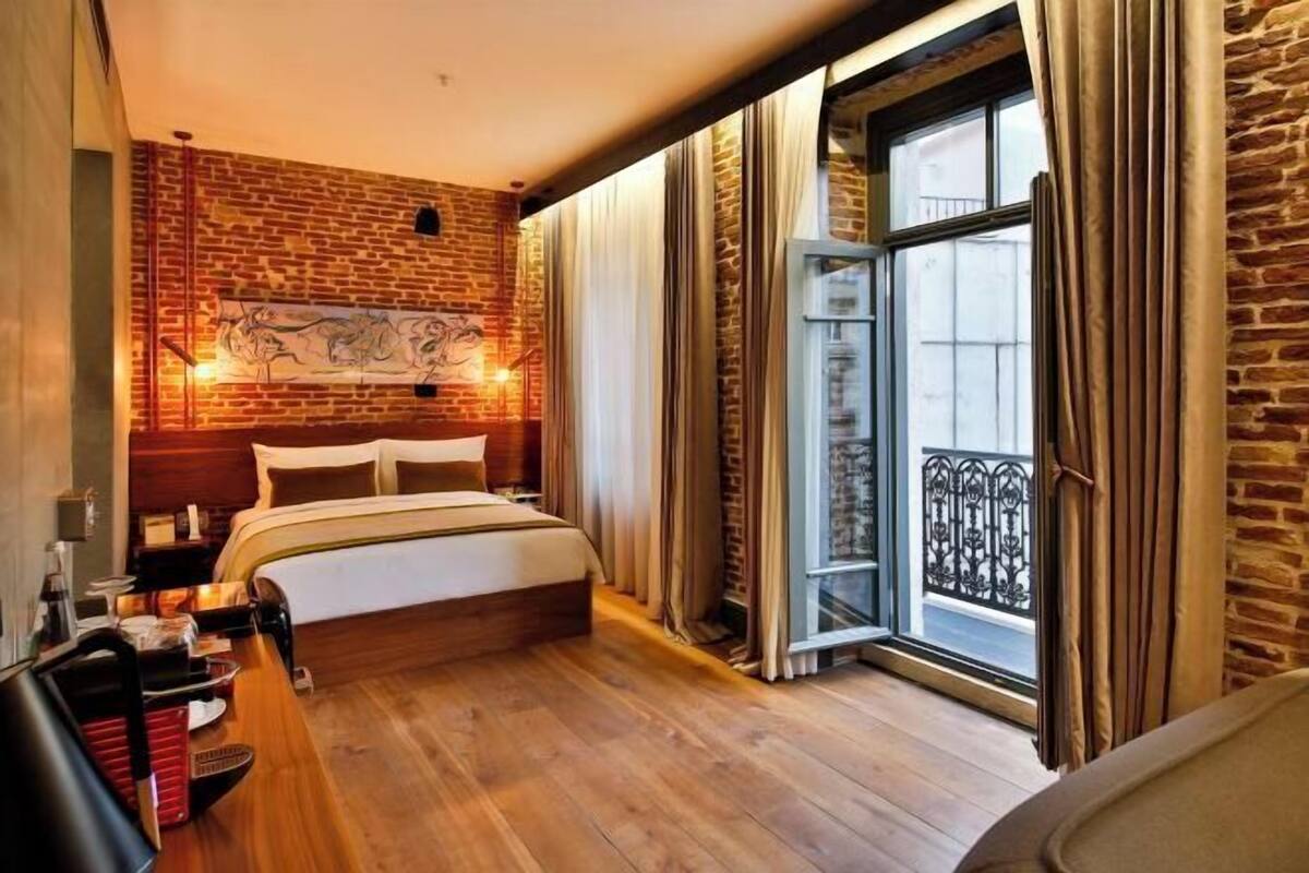 Una habitación de hotel con pared de ladrillo, una cama grande, suelo de madera, ventana con cortinas y balcón con barandilla de hierro forjado.