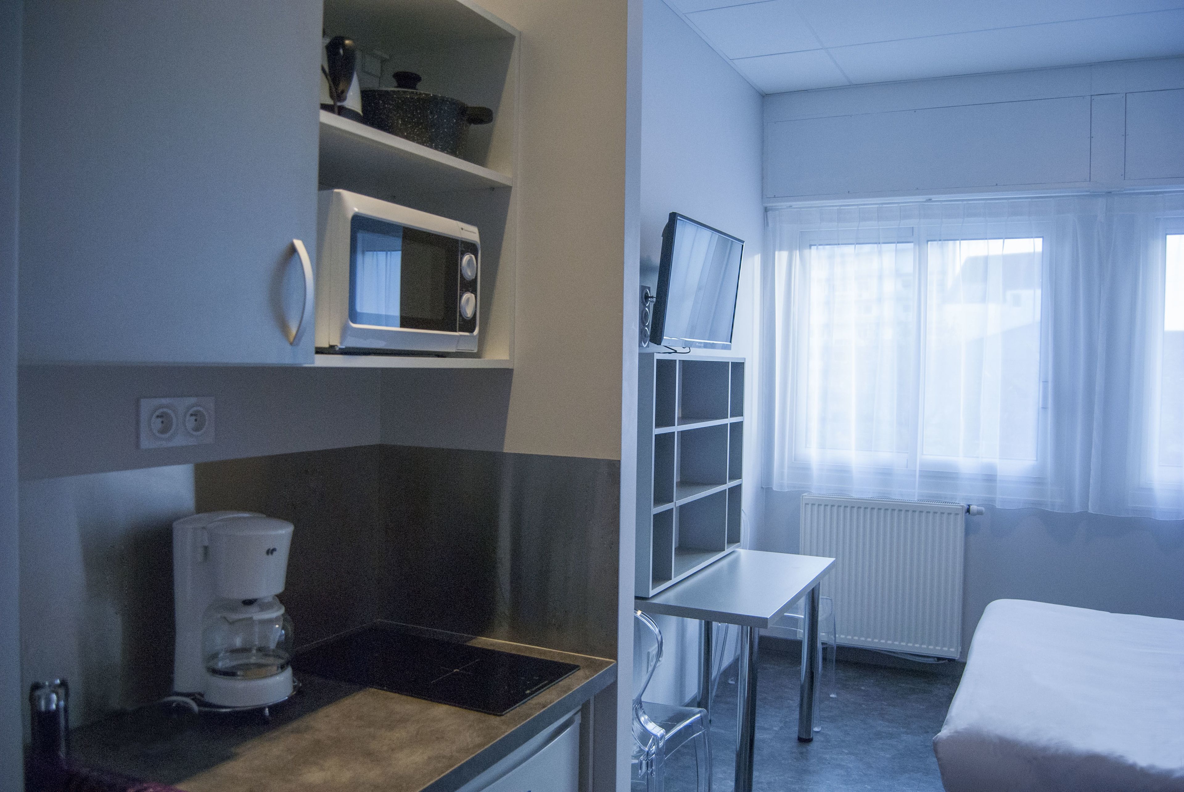 Een kleine kitchenette met een magnetron, koffiezetapparaat en een plank met keukengerei. Er is een kleine tafel en een stoel, en een raam met dunne gordijnen.