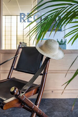A wooden chair with a straw hat placed on it, positioned in front of a window with a view of a building.