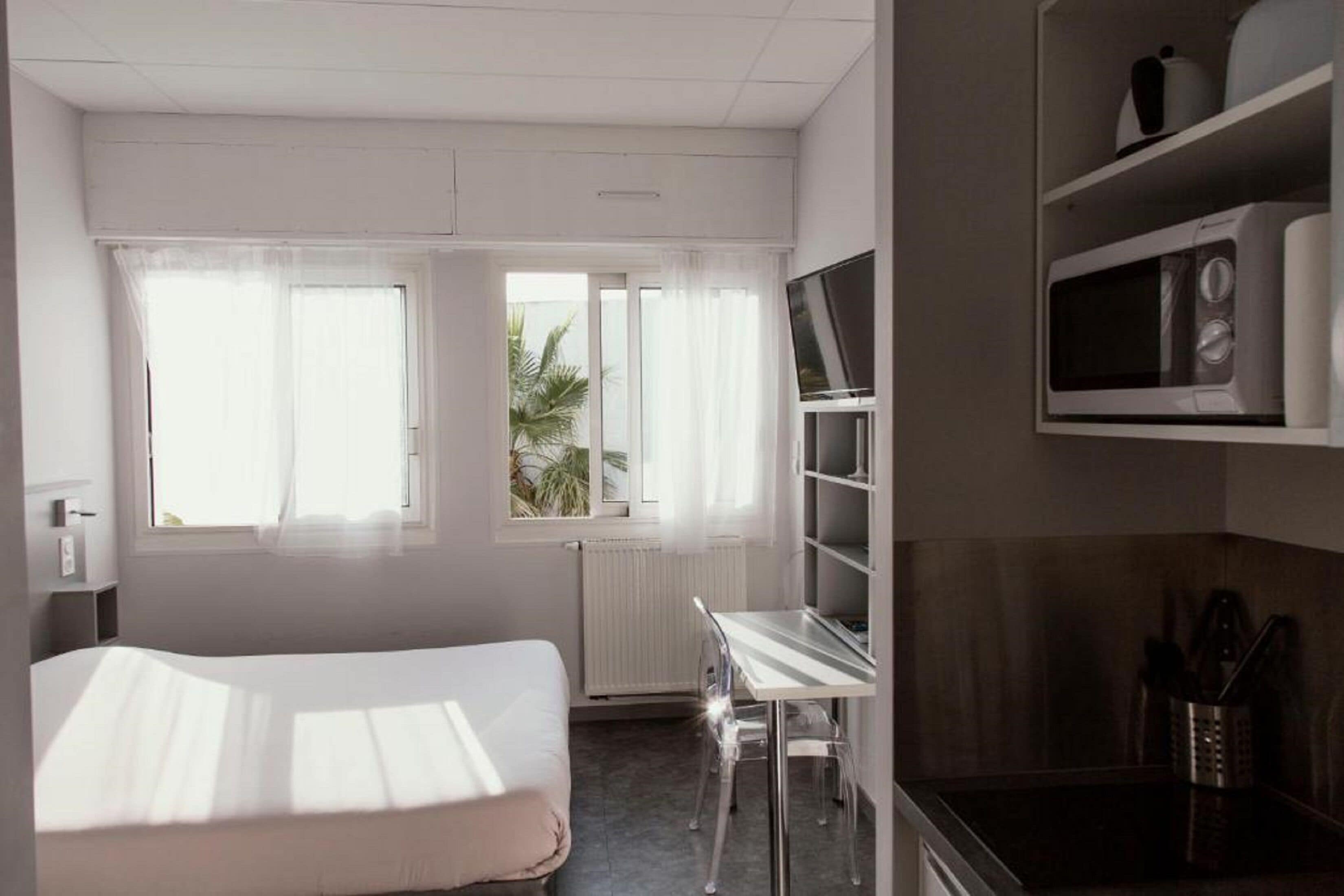 Quarto de hotel pequeno com cama, escrivaninha, micro-ondas e janela com cortinas.