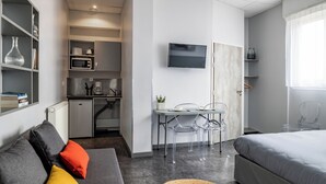 Kompakti hotellihuone, jossa on harmaa sohva, sänky, ruokapöytä tuoleineen, televisio ja pieni keittiö.