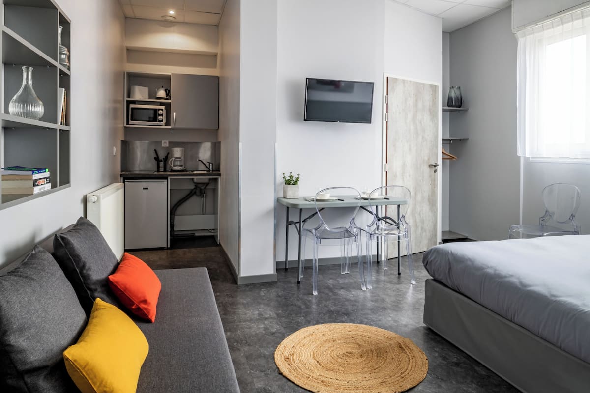 Een compacte hotelkamer met een grijze bank, een bed, een eettafel met stoelen, een televisie en een kitchenette.