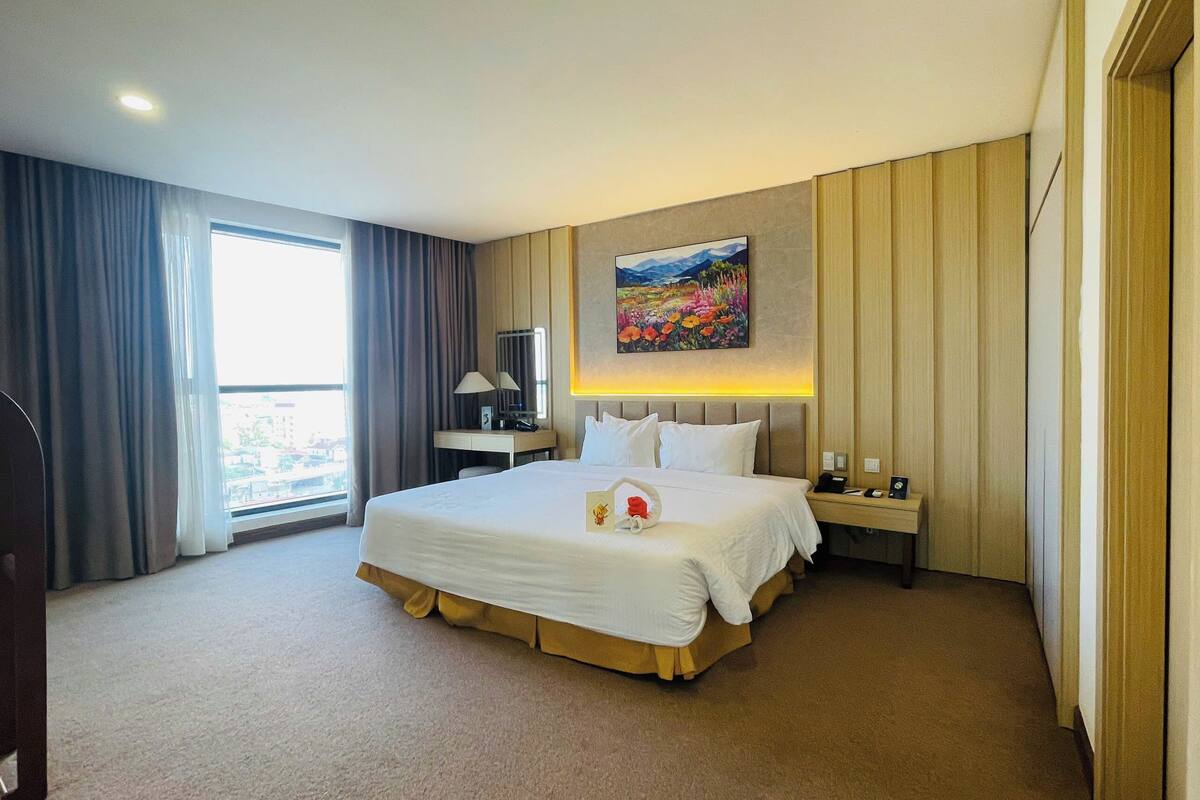 A hotel room with a large bed, a desk, a chair, a lamp, and a painting on the wall.