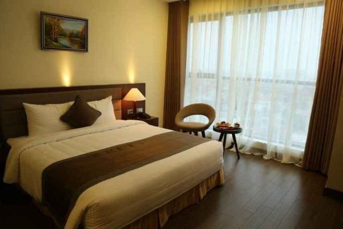 Een hotelkamer met een groot bed, een nachtkastje met een lamp, een stoel en uitzicht door dunne gordijnen.