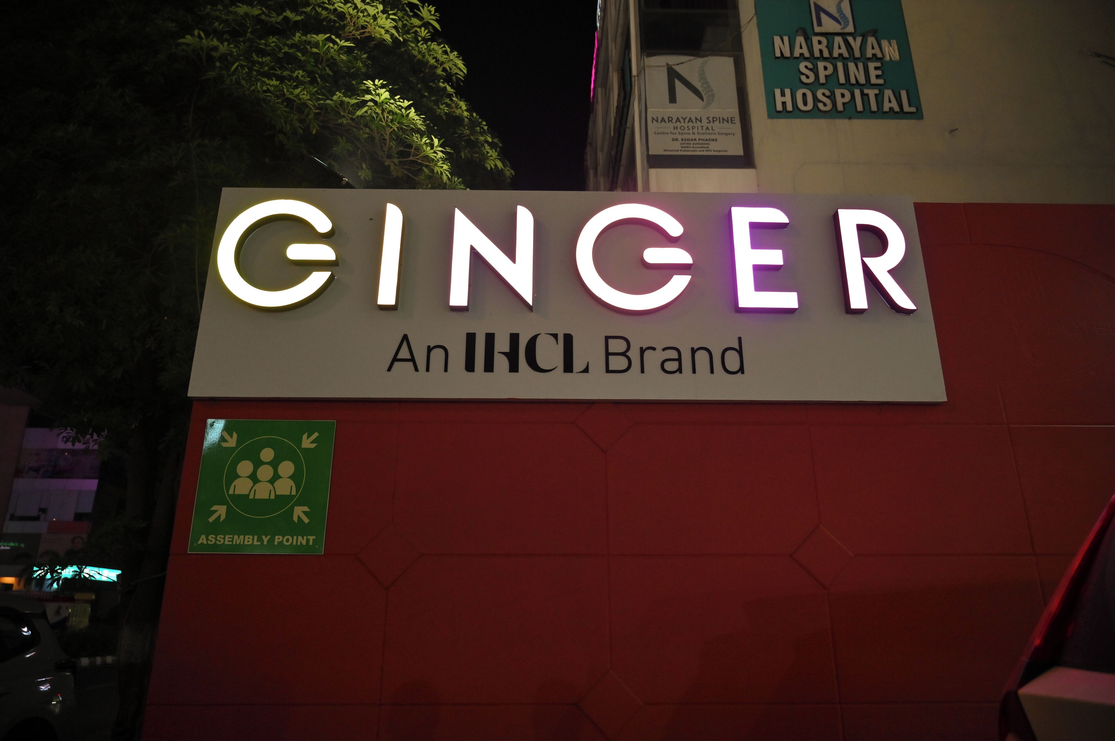 Señal de 'GINGER An IHCL Brand' con un símbolo de punto de encuentro y, al fondo, un letrero del Hospital de Columna Narayan.