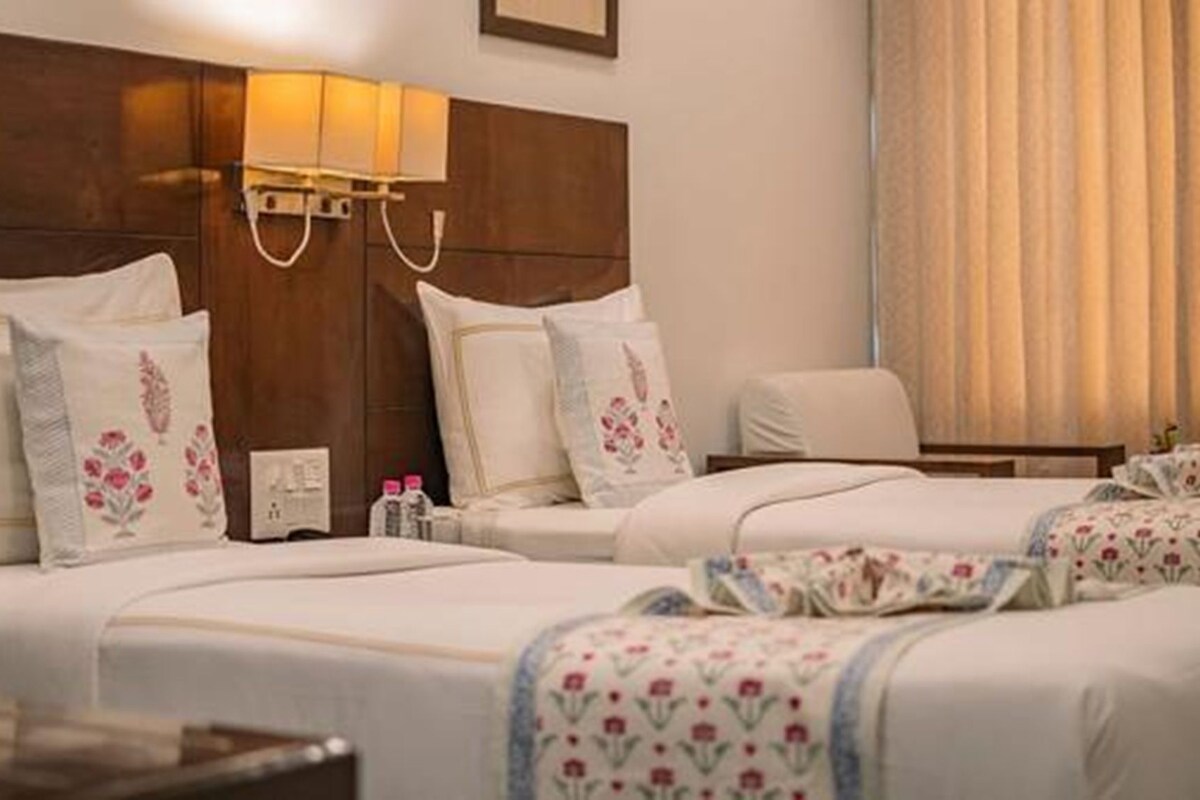 A hotel room with two beds, each with floral-patterned bedding and matching pillows. There is a bedside table with a lamp and a small clock. The room has a wooden headboard and a window with curtains.