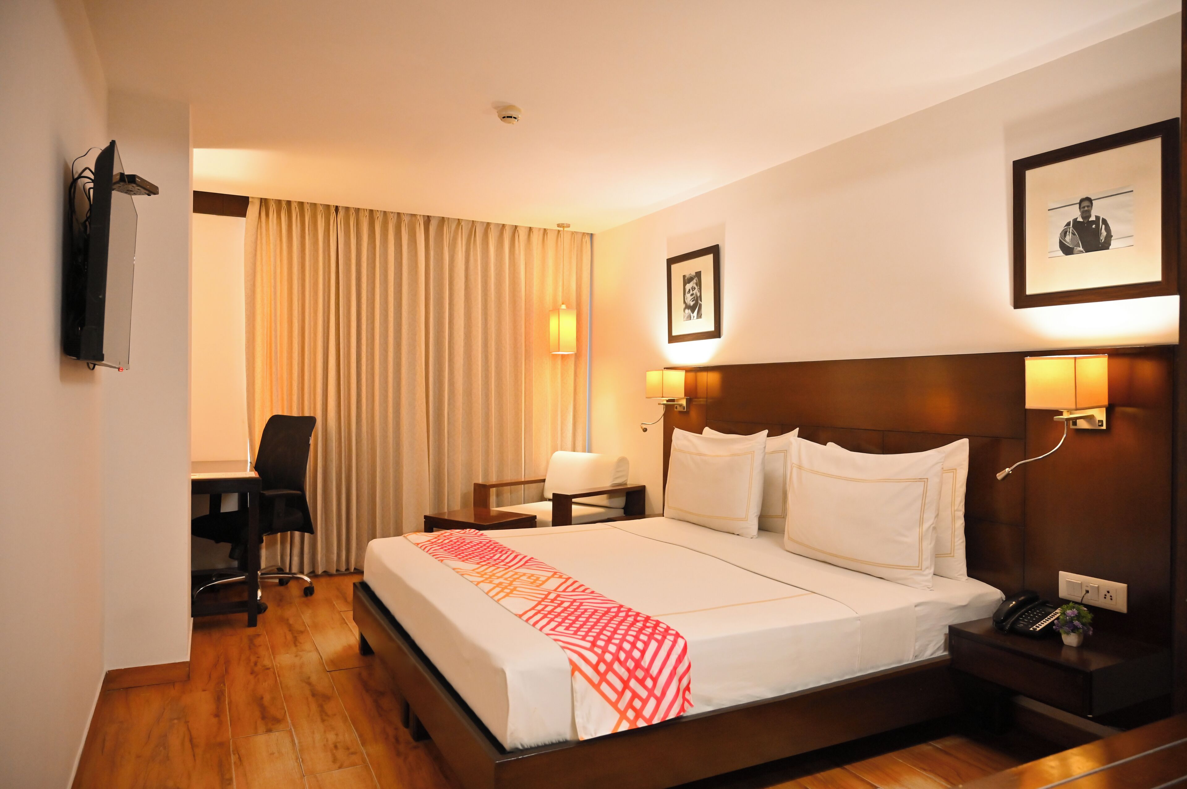 A hotel room with a bed, bedside lamps, a desk, a chair, and a framed picture on the wall.