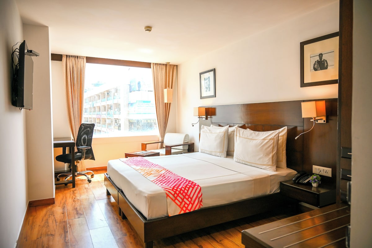 A hotel room with a large bed, a desk, a chair, a TV, a window with curtains, a framed picture, and a nightstand with a phone.