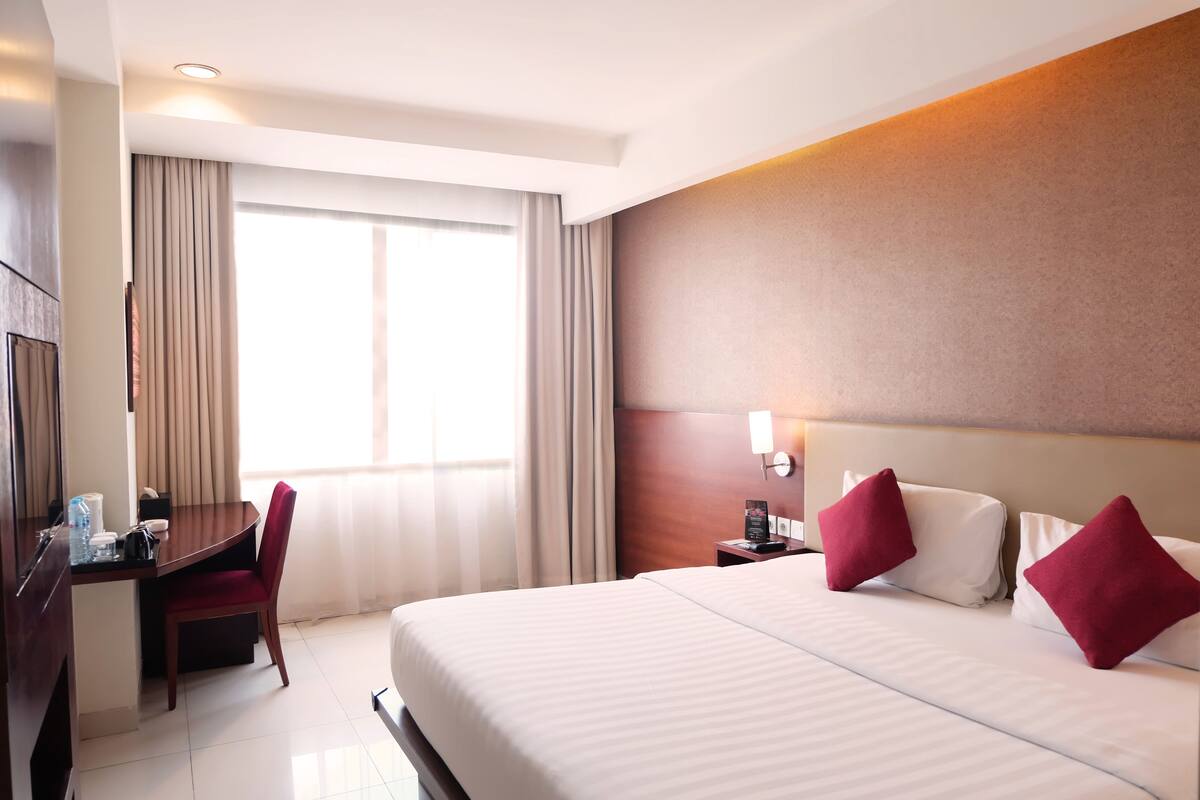 A hotel room with a bed, a desk, a chair, a nightstand, and a window with curtains.