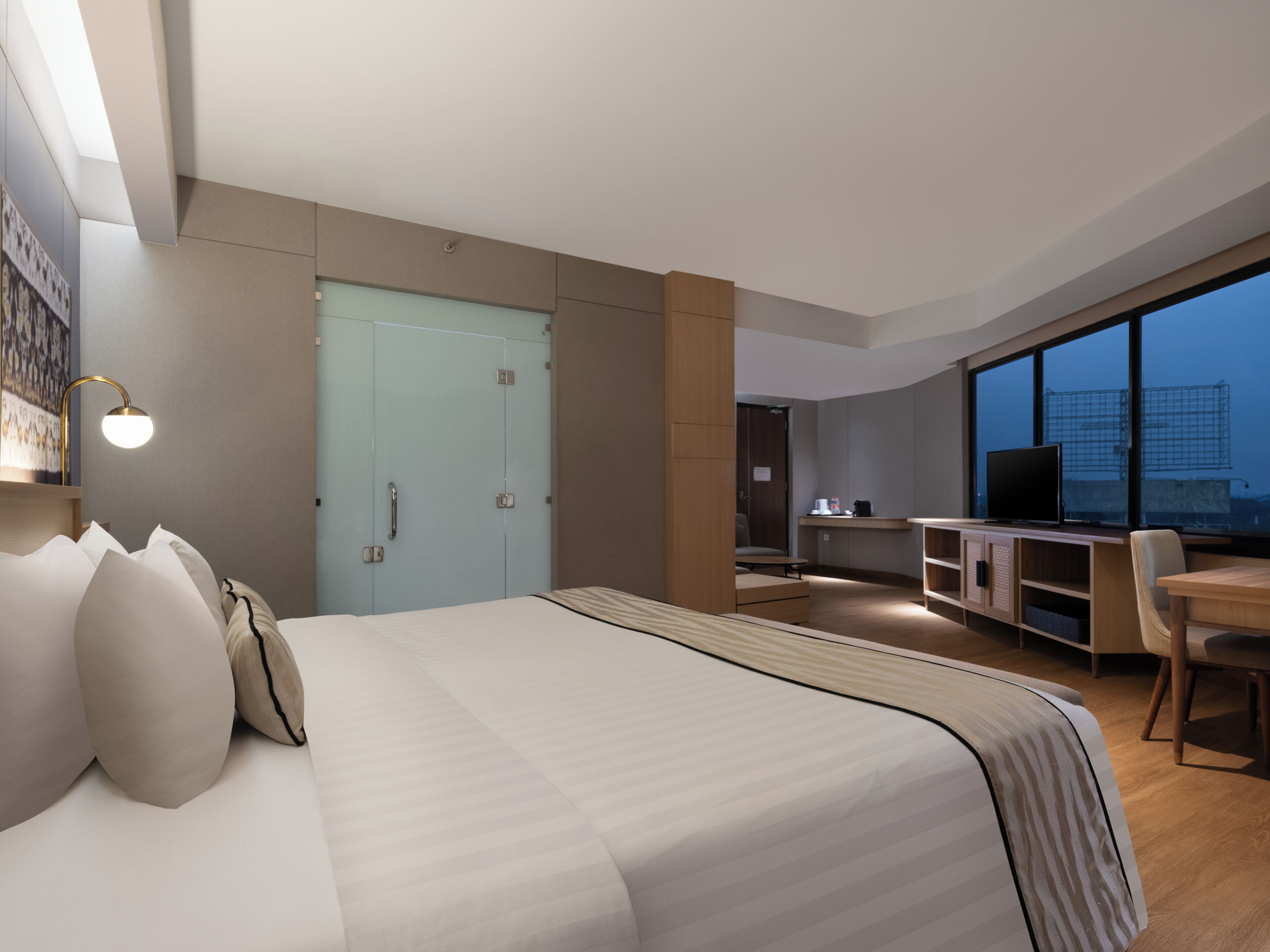 A modern hotel room with a large bed, a desk with a computer, a television, and a view of the cityscape through large windows.