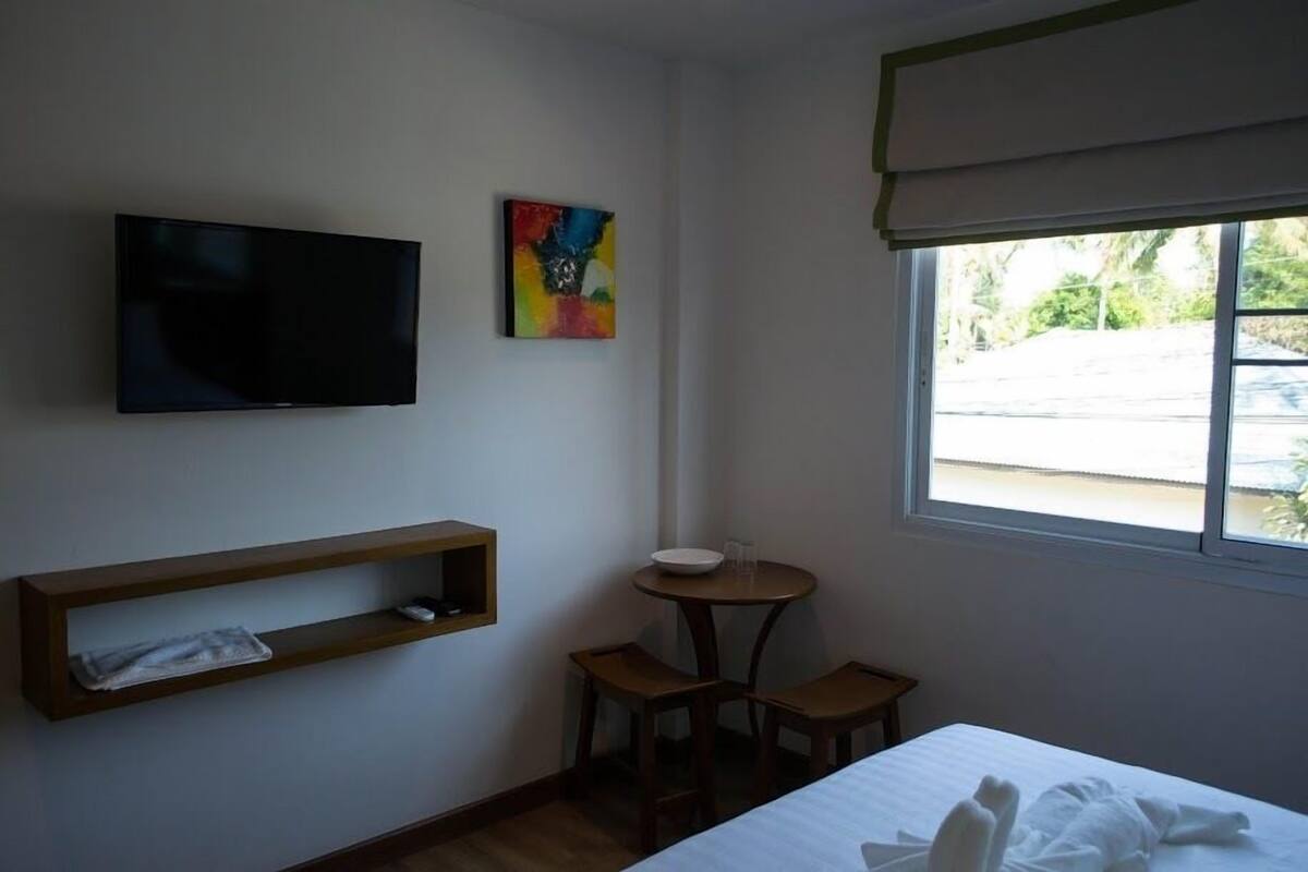 A hotel room with a bed, a TV mounted on the wall, a small table with chairs, and a window with a view of the outside.