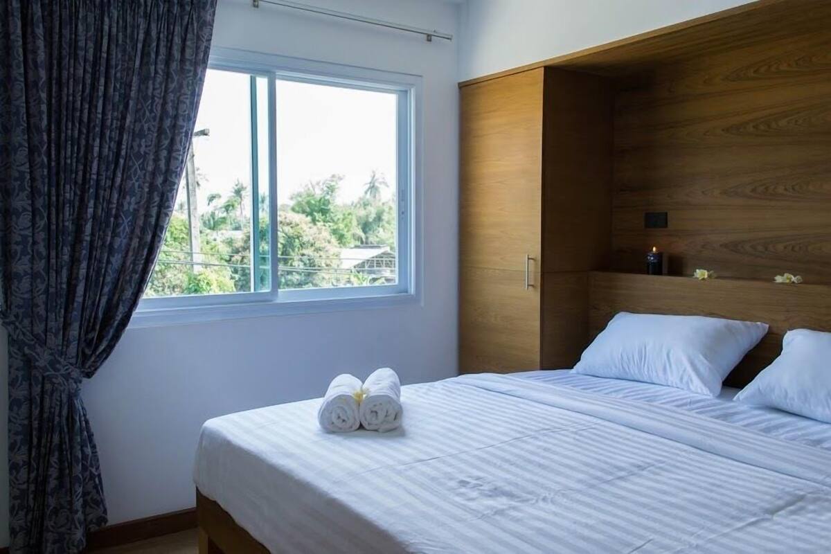 A neatly made bed with white linens and two rolled towels, a wooden wardrobe, and a window with a view of greenery outside.